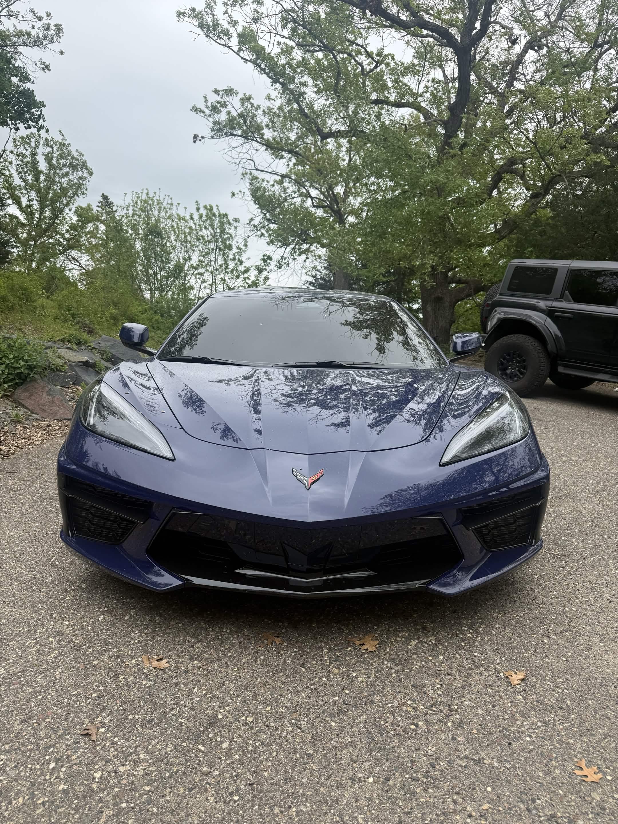 Front view of a dark blue Corvette C8 parked on asphalt surrounded by green trees.