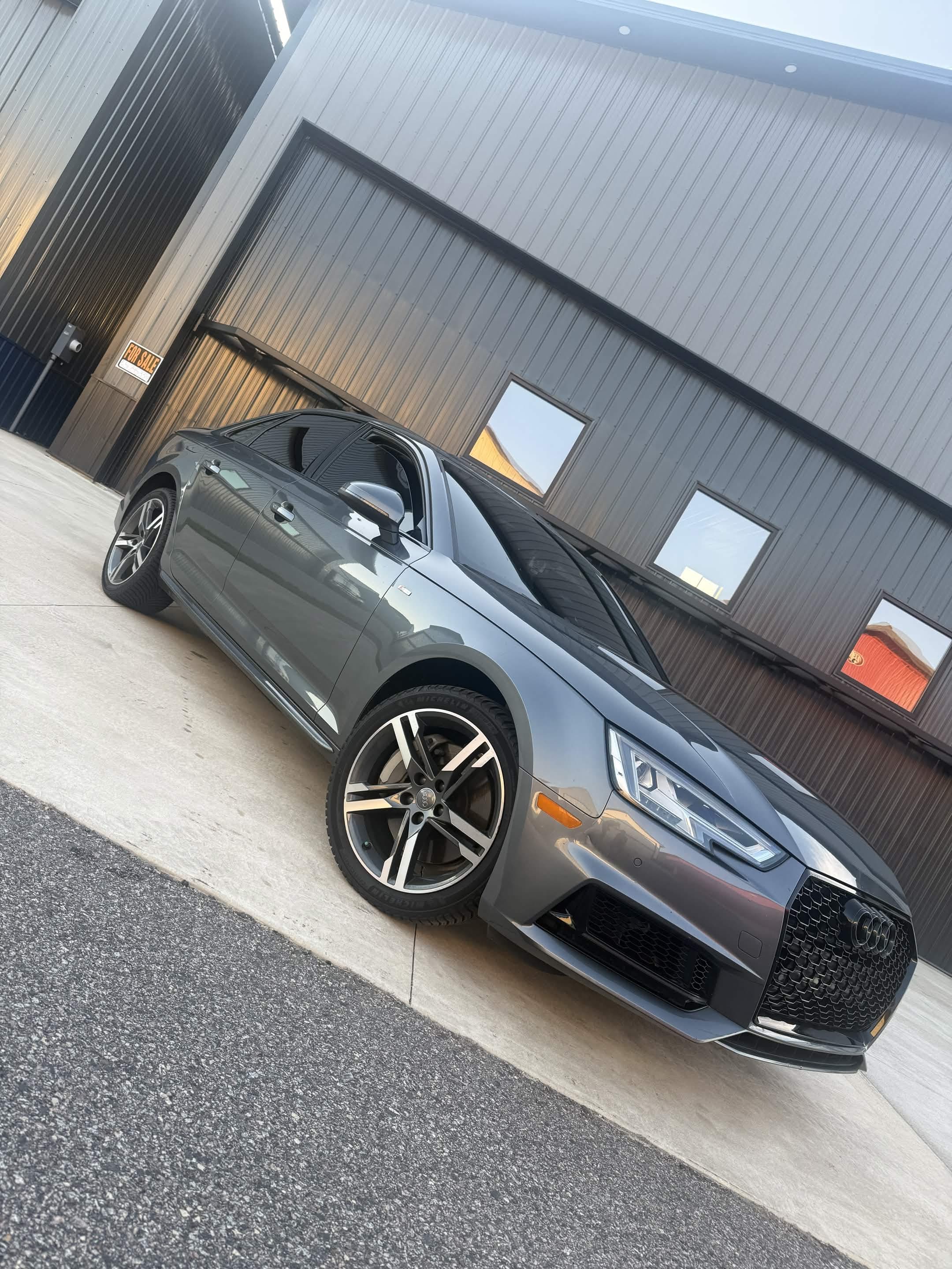 Sleek gray Audi sedan parked in front of a modern gray corrugated metal industrial building.